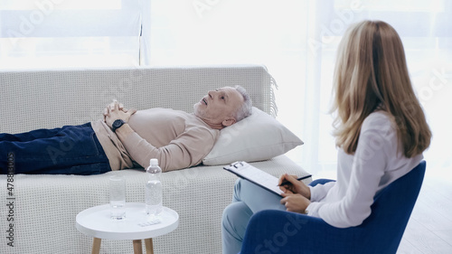 blonde psychotherapist holding pen near clipboard and listening middle aged man lying on couch in consulting room © LIGHTFIELD STUDIOS