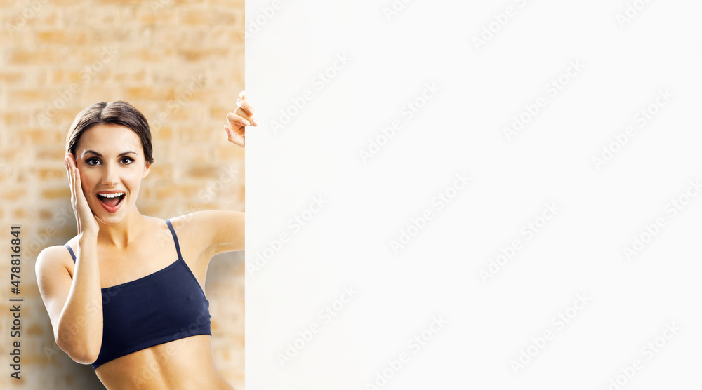 Photo of excited beautiful brunette woman in fitness wear showing signboard with copy space. Fit girl with happy wide open mouth, peek out mock up white placard, bricks wall background. - obrazy, fototapety, plakaty 