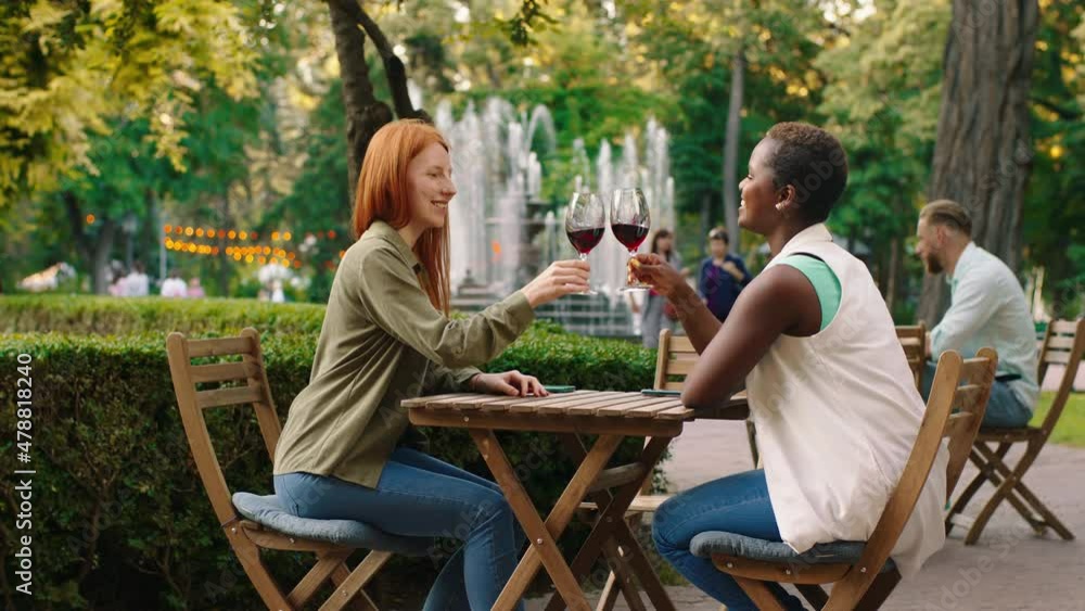 An angelic looking woman with ginger long hair is sharing and cheering some wine with her African friend, they are sitting at a table and surround by the park An angelic looking woman with ginger long hair is sharing and cheering some wine with her African friend, they are sitting at a table and surround by the park