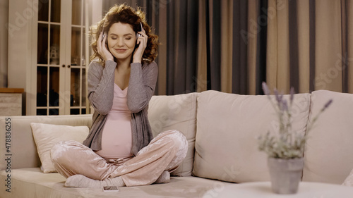Cheerful pregnant woman in headphones sitting near smartphone on couch
