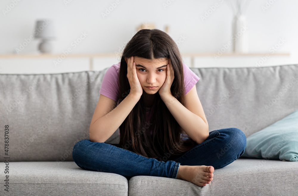 Upset Indian teenage girl sitting on couch, feeling bored, suffering from lack of communication, being trapped at home Upset Indian teenage girl sitting on couch, feeling bored, suffering from lack of communication, being trapped at home