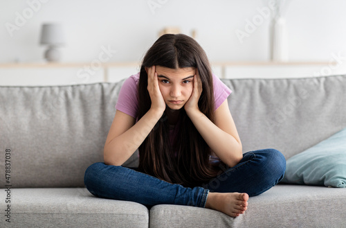 Upset Indian teenage girl sitting on couch, feeling bored, suffering from lack of communication, being trapped at home