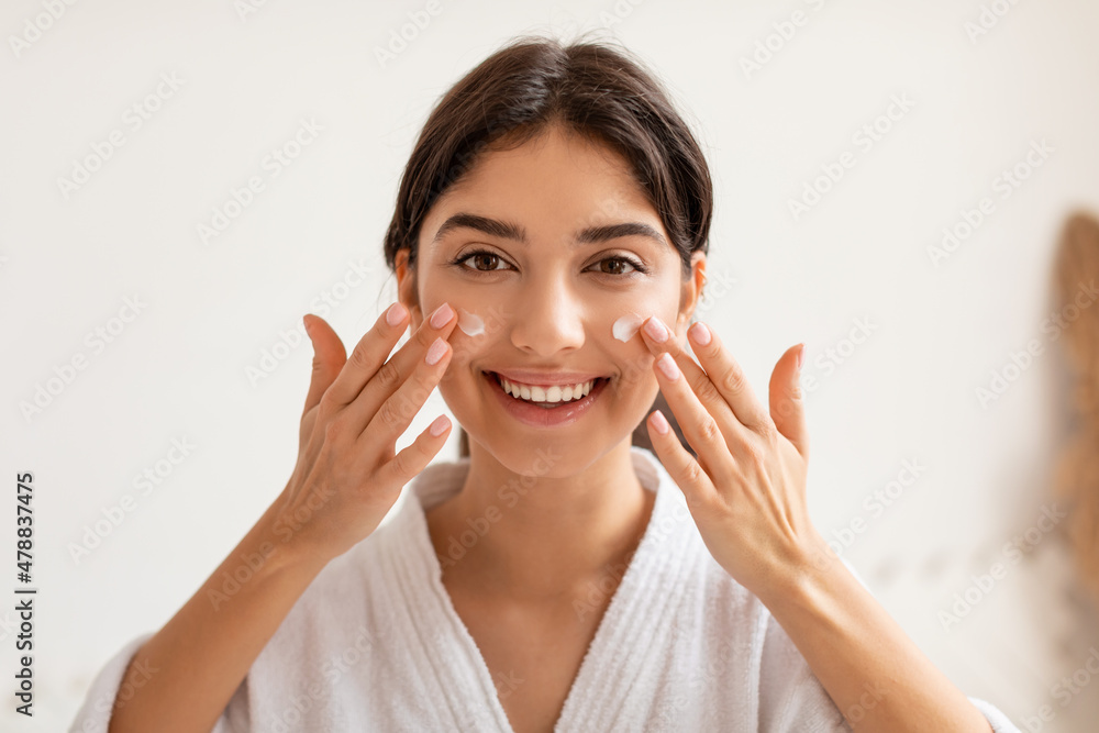 Cheerful Woman Applying Facial Cream Moisturizing Skin Posing In Bathroom