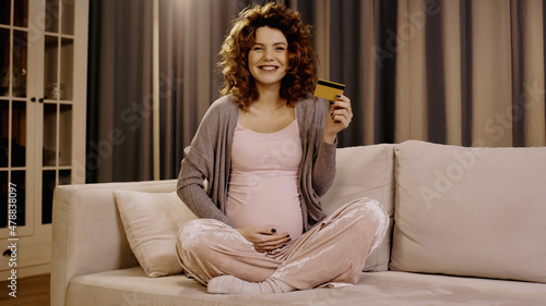 Positive pregnant woman holding credit card and smiling at camera at home