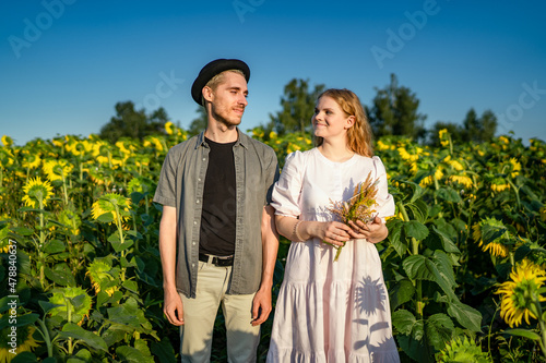 young pretty blonde caucasian girl with curly hair in a light dress in a sunflower field on a sunny day smiling, posing on the phone camera with a cute guy in a black hat