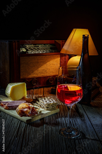 Glass and bottle of red wine. Plate with toast, ham, bacon, cheese. Old radio and lampshade. Αtmosphere of warm low light.