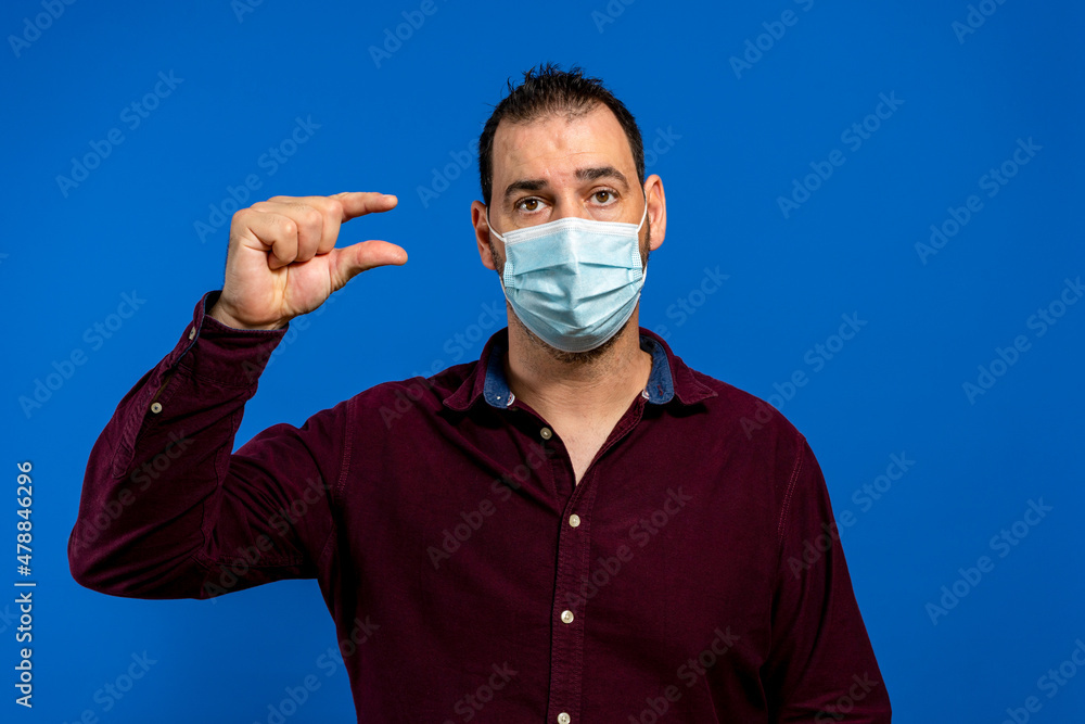 Young caucasian man with short hair wearing medical mask standing over isolated blue background smiling and confident gesturing with hand doing small size sign with fingers looking and the camera Young caucasian man with short hair wearing medical mask standing over isolated blue background smiling and confident gesturing with hand doing small size sign with fingers looking and the camera