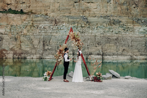 Guy and girl on their wedding day stand near a boho-style wedding decor, mountains, a lake and stones. A girl in a dress and a white hat and a guy in a black suit and a white shirt