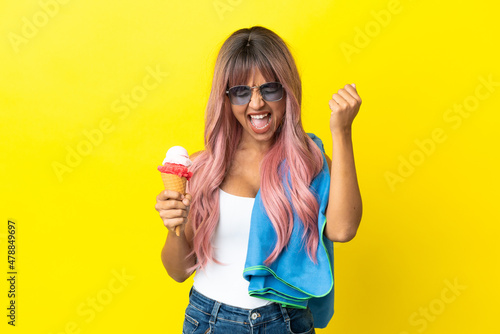 Young mixed race woman with pink hair holding ice cream isolated on yellow background celebrating a victory