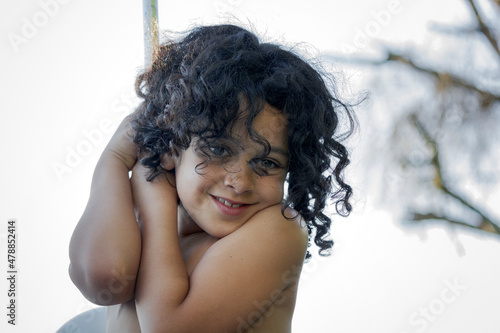 Niña de cabello negro rizado feliz agarrada a una barra en verano sonriendo y mirando a la cámara.