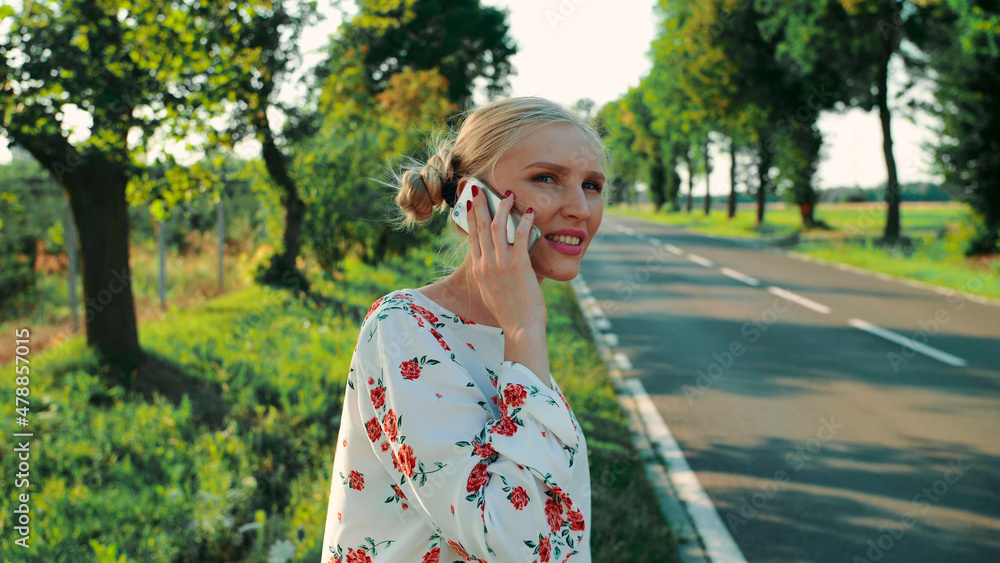 Woman speaking on phone while hitchhiking. Side view of lovely young female talking on smartphone and waiting for car while standing on roadside during trip through countryside.