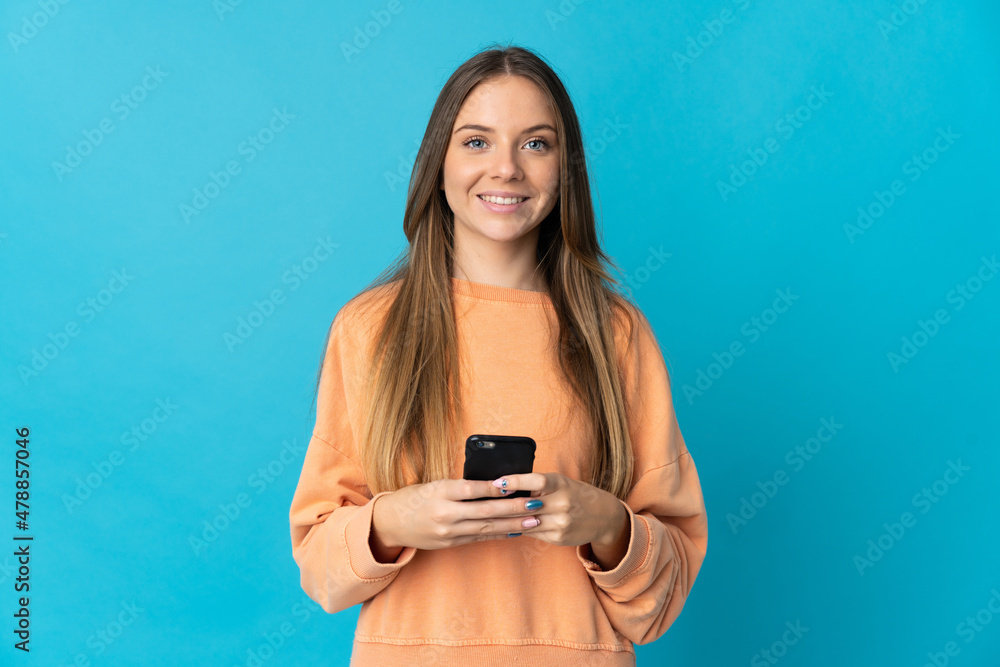 Young Lithuanian woman isolated on blue background looking at the camera and smiling while using the mobile Young Lithuanian woman isolated on blue background looking at the camera and smiling while using the mobile