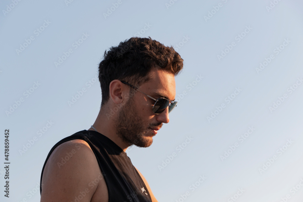Obraz premium Fit, handsome boy with sun glasses. He is on summer vacation. Thoughtful face. He is looking down. Sunlight. Cover boy. Copy and text space. 