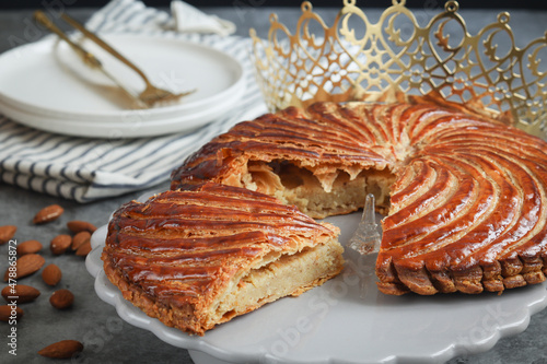 A traditional French galette des rois on a plate with crown, with a cut piece and its fève in a middle. Made puff pastry and creamy almond filling roll in circle shape. It's usually served on Epiphany photo