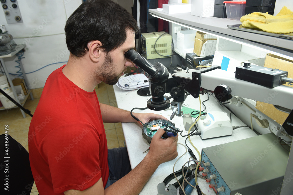 A young man in the workshop examines the microcircuit of an electrical device through a microscope, performing electrical soldering of equipment A young man in the workshop examines the microcircuit of an electrical device through a microscope, performing electrical soldering of equipment