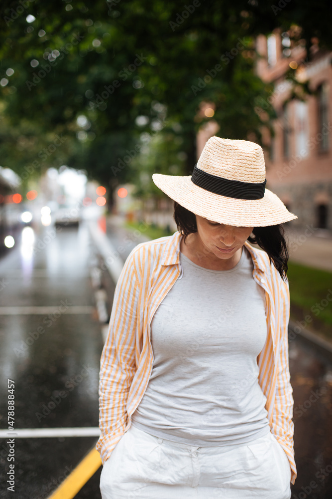 Hidden face portrait of woman walking in the middle of the street. Female in summer rain. City lifestyle fashion. Woman wearing summer clothes and hat.