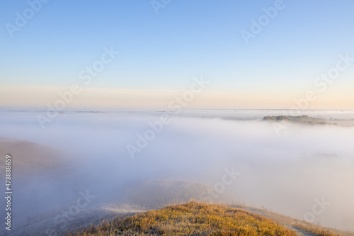 Fototapeta Naklejka Na Ścianę i Meble -  Autumn landscape in the early morning. Fog-covered expanses through which the first rays of the rising sun pass. Trees and hills in the fog. Dawn on a cold autumn morning.
