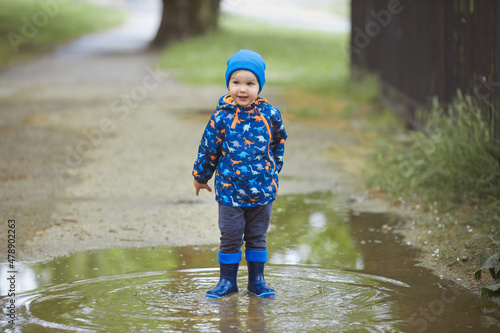 Fototapeta Naklejka Na Ścianę i Meble -  Portrait of happy toddler boy 2 years old in a waterproof raincoat and blue rubber rain boots in early spring have fun, playing and running through the puddles in the city street. 