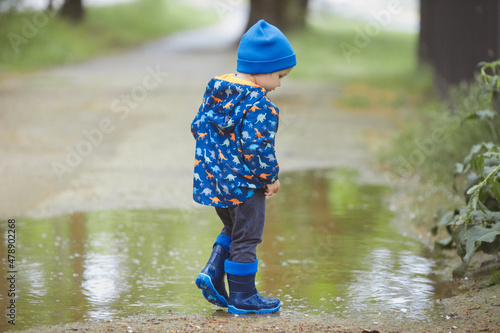 Portrait of happy toddler boy 2 years old in a waterproof raincoat and blue rubber rain boots in early spring have fun, playing and running through the puddles in the city street. 