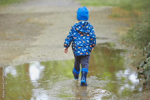 Faceless funny toddler boy 2 years old from behind in a waterproof jacket and blue rubber boots in early spring have fun and running through the puddles after the rain in the city. 