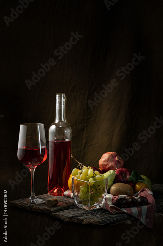 Red wine glass and bottle. Cheese, ham, grapes, pomegranate, kiwi, mandarin. Wooden table.