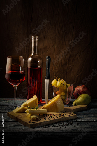 Red wine glass and bottle. Cheese, ham, grapes, pomegranate, kiwi, mandarin. Wooden table.
