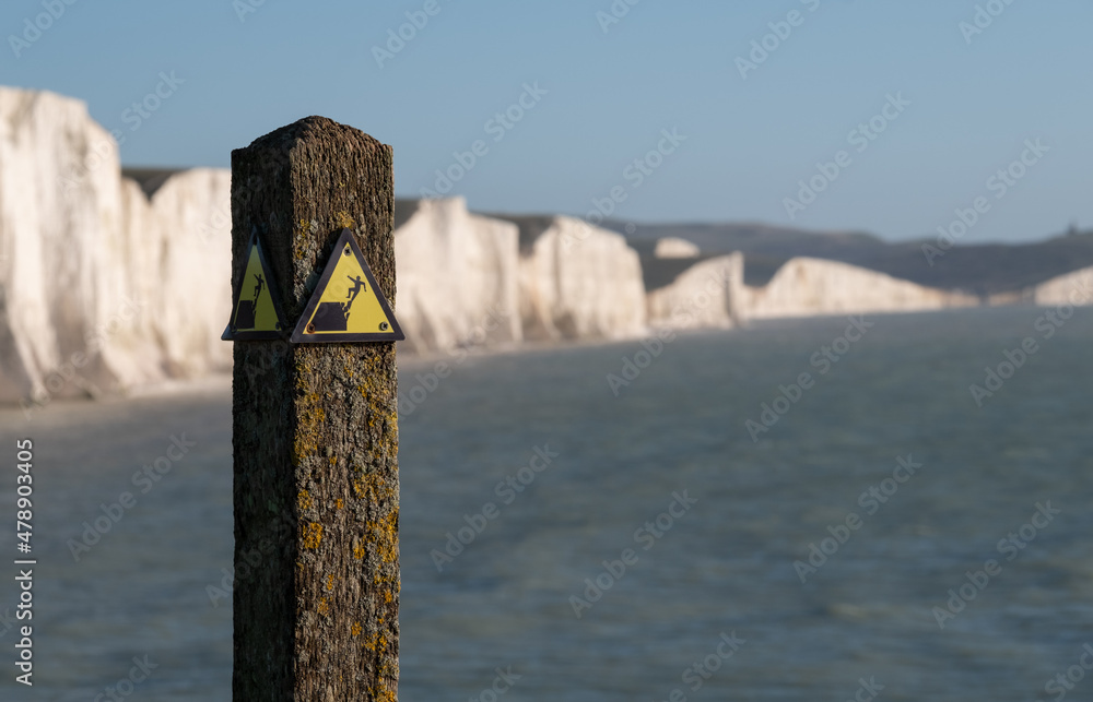 Yellow and black danger sign warning of erosion at the cliff edge. In the background Seven Sisters chalk cliffs at Seaford in East Sussex on the south coast of England UK. Photographed at Hope Gap. Yellow and black danger sign warning of erosion at the cliff edge. In the background Seven Sisters chalk cliffs at Seaford in East Sussex on the south coast of England UK. Photographed at Hope Gap.