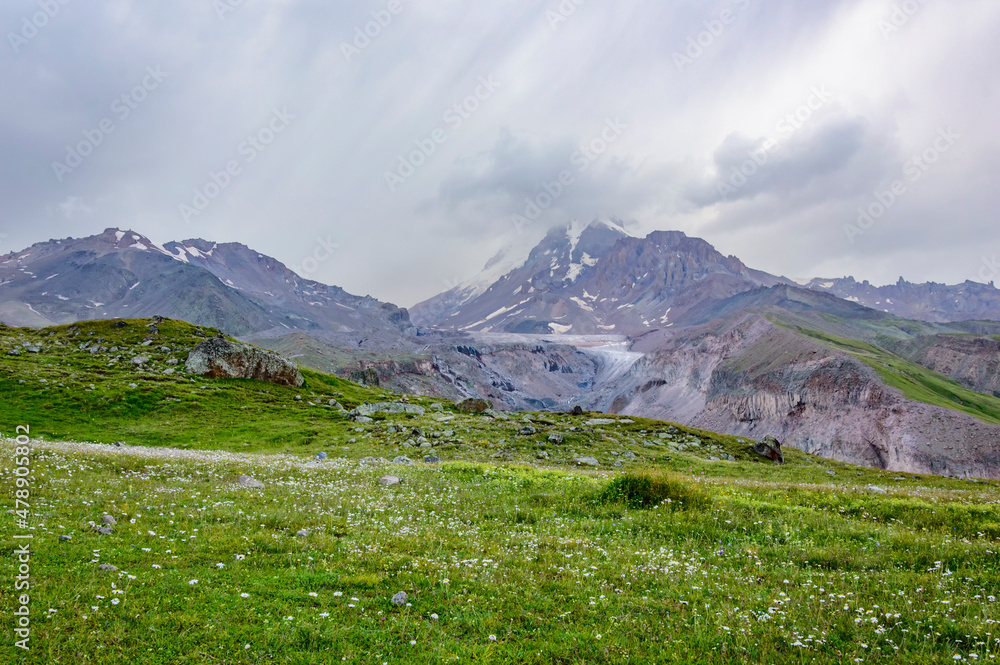 Grey thunder sky with clouds above mount Kazbek in Georgia, grey and brown slopes with snow, green hills with grass and flowers Grey thunder sky with clouds above mount Kazbek in Georgia, grey and brown slopes with snow, green hills with grass and flowers