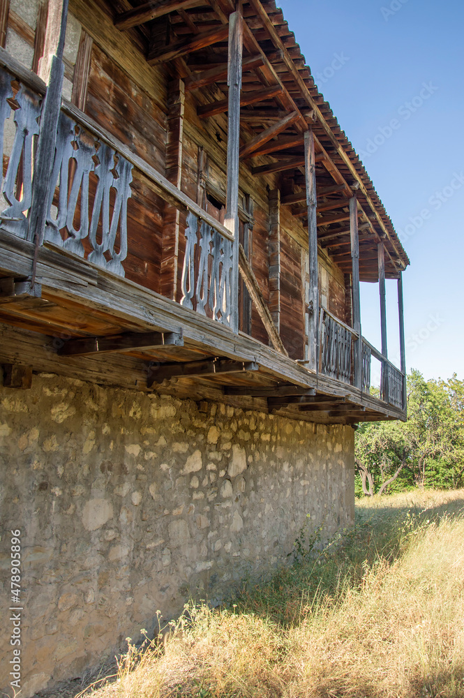 Abandoned traditional georgian house in Tbilisi. Stone and wooden walls,  ruined wooden balcony, dry grass, green trees, blue sky