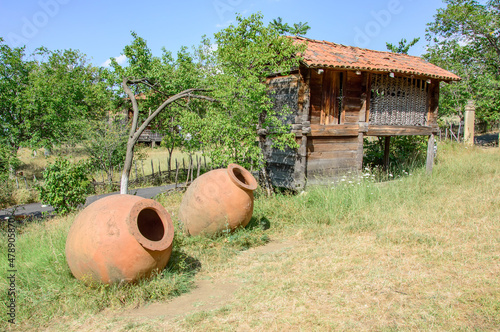 Traditional georgian house and two big kvevris for vine in Tbilisi  open-air ethnographical museum. Wooden walls, tiled roof, dry grass, green trees and bushes, blue sky photo