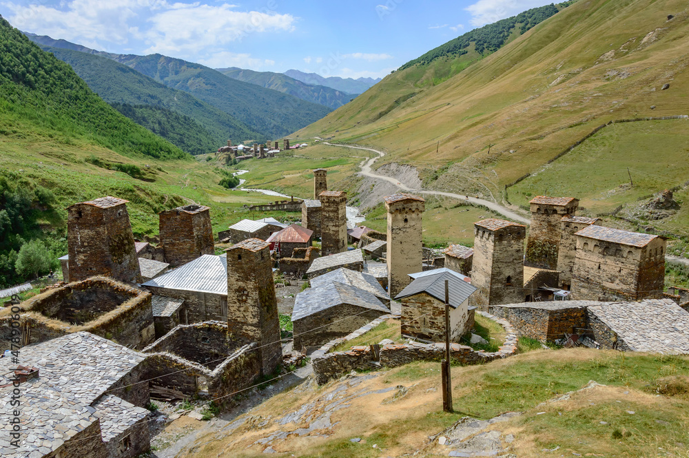 Hilltop view of Ushguli fortified village in Svanetia, Georgia. Grey, brown and biege medieval stone towers and houses, green and yellow dry grass, road, green mountains and blue sky with clouds
