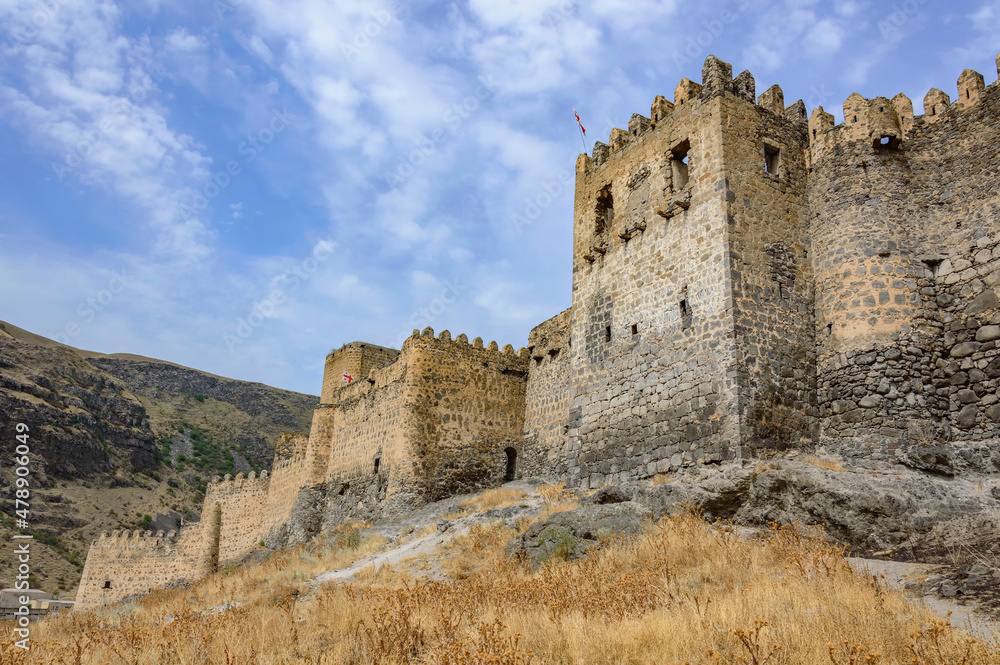 Khertvisi fortress in Georgia, Caucasus. Ancient weathered medieval grey and biege stone walls and towers, grey cliff, blue sky with clouds, dry yellow grass