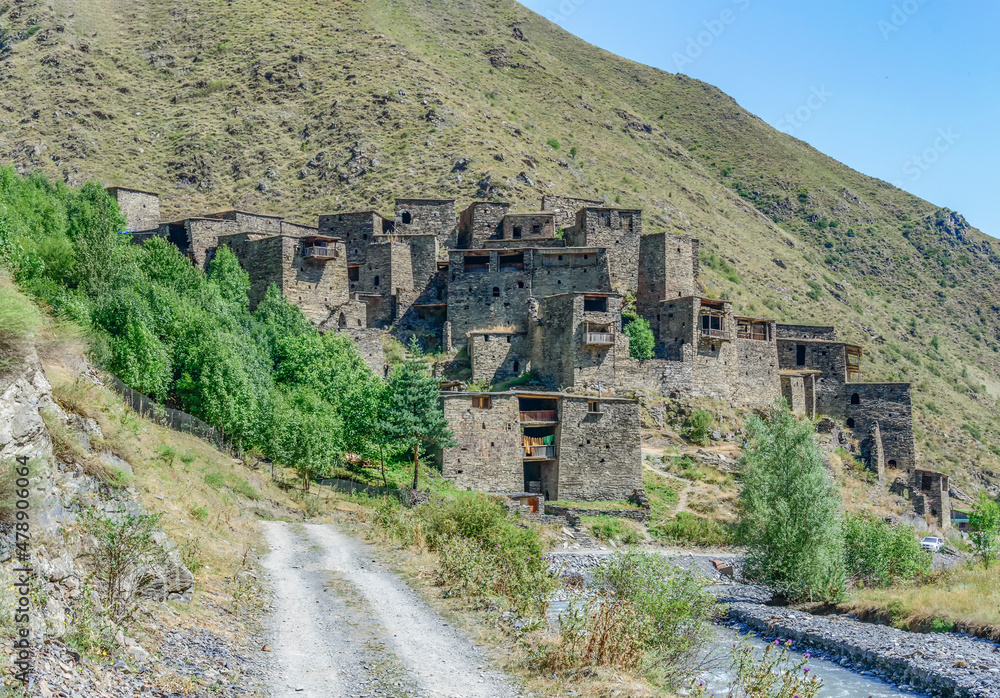 The Fortified Village of Shatili, Khevsureti, Georgia. Abandoned towers and houses, brown and grey old stone walls, green trees and dry grass on mountain slopes, blue sky with clouds, road and river