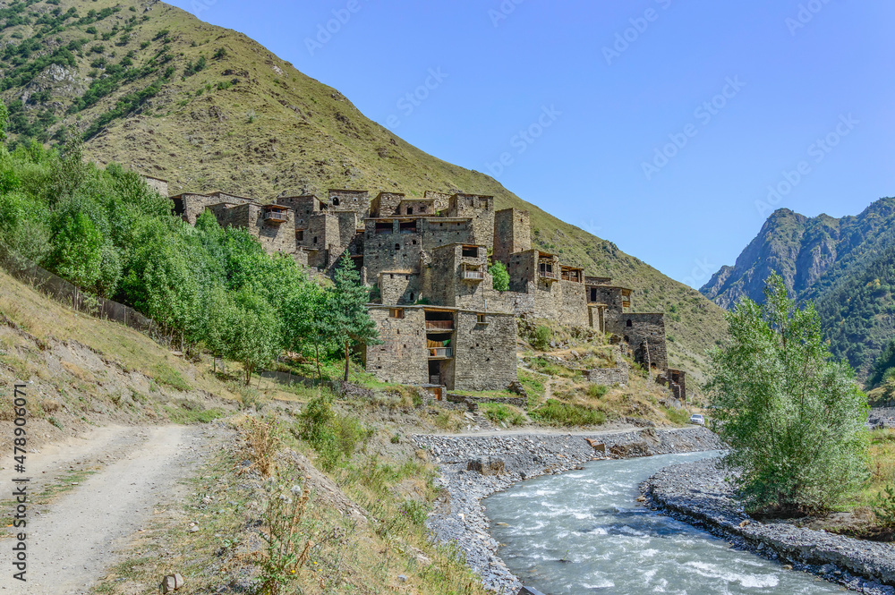 The Fortified Village of Shatili, Khevsureti, Georgia. Abandoned towers and houses, brown and grey old stone walls, green trees and dry grass on mountain slopes, blue sky with clouds, road and river