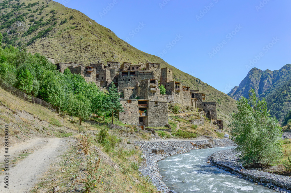 Fototapeta premium The Fortified Village of Shatili, Khevsureti, Georgia. Abandoned towers and houses, brown and grey old stone walls, green trees and dry grass on mountain slopes, blue sky with clouds, road and river