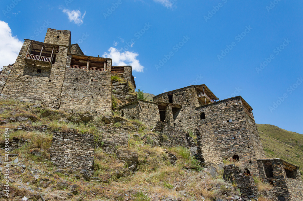 The Fortified Village of Shatili, Khevsureti, Georgia. Abandoned towers and houses, brown and grey old mossy stone walls, green trees and dry grass on mountain slopes, blue sky with clouds.