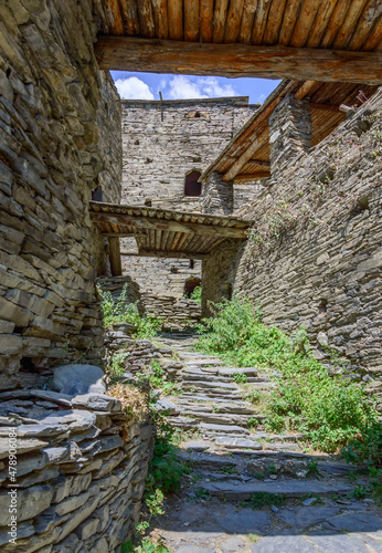 Narrow street in the Fortified Village of Shatili, Khevsureti, Georgia. Abandoned houses, grey old stone walls, green bushes and grass, stone stairs, blue sky with clouds.
