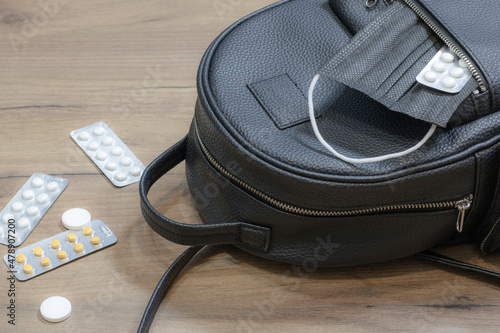 A woman's bag and a medical mask with medicines in an open pocket, medicines are scattered on the table next to them.