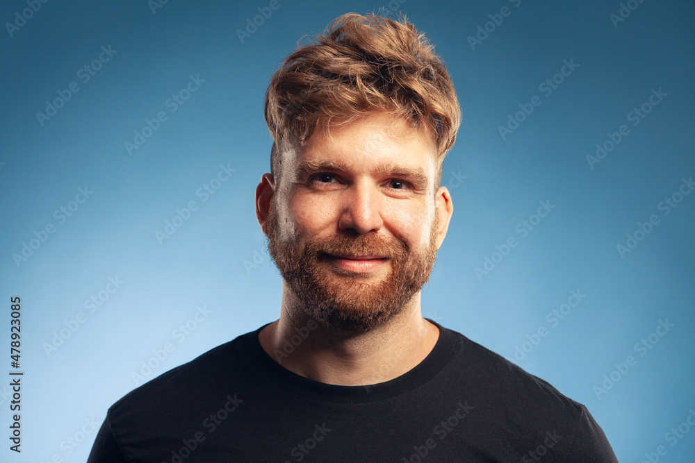 Fototapeta premium Male beauty concept. Emotive portrait of cute charismatic active 30-year-old man posing over blue background. Perfect haircut. Hipster style. Close up. Text space. Studio head shot