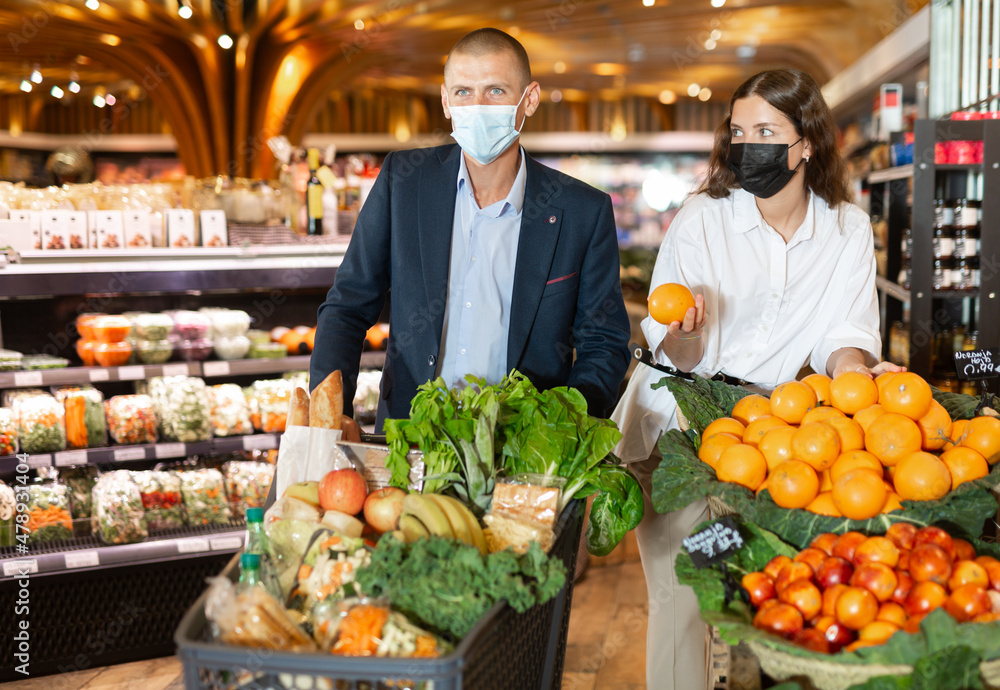Portrait of young woman and man in face mask for disease protection doing shopping at vegetables department at supermarket during coronavirus pandemic