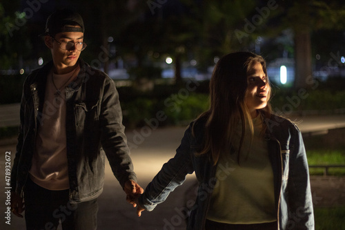 young latin couple walking in the park at night holding hands, she is ahead of him and he looks at her hand with affection, the night lights illuminate their faces.