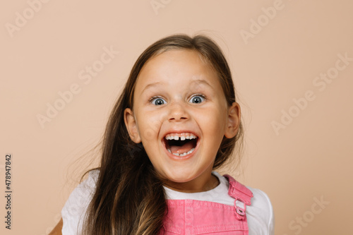 Close-up face kid with shining eyes and overexcited smile with teeth, opened mouth and raised eyebrows looking at camera wearing bright pink jumpsuit and white t-shirt on beige background. photo