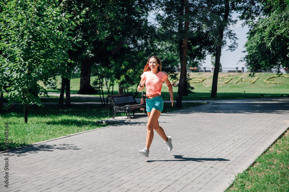 running smiling young sporty woman with watch and flowing hair in park in pink t-shirt, green shorts and white trainers on background of green trees and grass in front of bench and inscription 