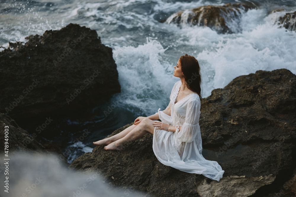 A woman in a white dress barefoot sits on a stone on a cliff top view unaltered