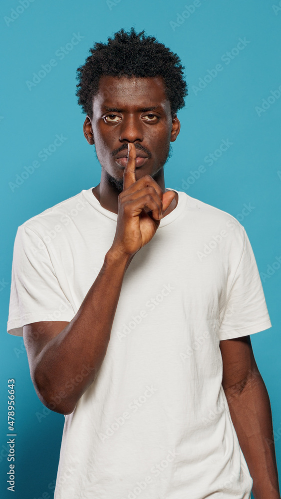 Young man doing silence gesture with finger on mouth in studio. Confident person asking for secrecy and quiet, using forefinger to make censorship expression over isolated background. Young man doing silence gesture with finger on mouth in studio. Confident person asking for secrecy and quiet, using forefinger to make censorship expression over isolated background.