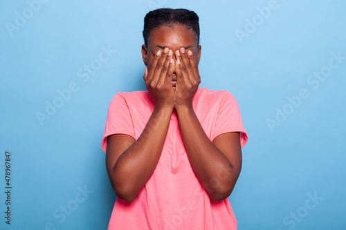 Portrait of scared stressed african american young woman covering face with her hands during photo shotting having social anxiety standing in studio with blue background. Terrified introvert model