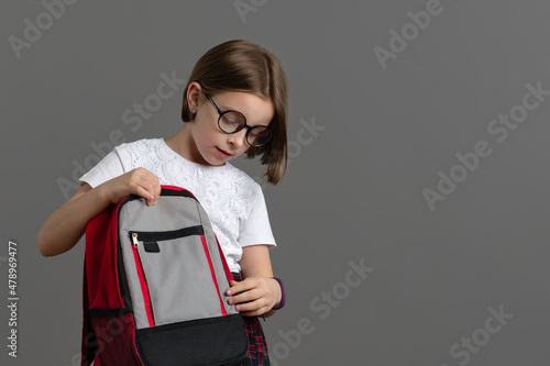 Smart Little Girl 9s Wearing school uniform and in stylish round glasses with book bag on grey background indoor. Clever school child checks school backpack. Educational concept for school photo