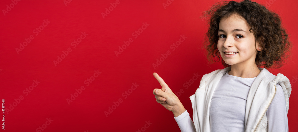 One small caucasian girl ten years old with curly hair front view portrait close up standing in front of red background looking to the camera pointing finger to copy space smiling happy and joy One small caucasian girl ten years old with curly hair front view portrait close up standing in front of red background looking to the camera pointing finger to copy space smiling happy and joy