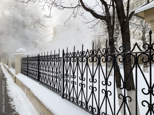 Sidewalk and iron fence in winter with steam in the background. White steam from a burst hot water pipe in winter.