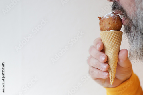 Man with grey beard eats horn with fried donut with sugar powder like ice-cream.